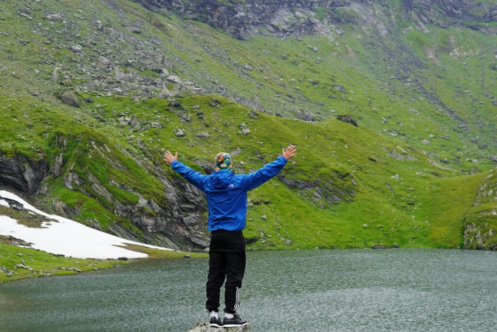 Imaginea prezintă o persoană cu brațele deschise, stând pe un piatră lângă un lac, înconjurată de dealuri verzi și pete de zăpadă. Această scenă de natură se asociază cu "Cum alegi influencerii potriviți în România fără să arunci bugetul pe geam", sugerând o abordare rațională și în harmonie cu medioanele naturale pentru a alege influențeri, evitând cheltuielile inutile.