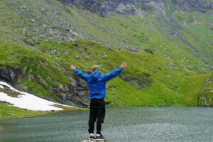Imaginea prezintă o persoană cu brațele deschise, stând pe un piatră lângă un lac, înconjurată de dealuri verzi și pete de zăpadă. Această scenă de natură se asociază cu &ldquo;Cum alegi influencerii potriviți în România fără să arunci bugetul pe geam&rdquo;, sugerând o abordare rațională și în harmonie cu medioanele naturale pentru a alege influențeri, evitând cheltuielile inutile.
