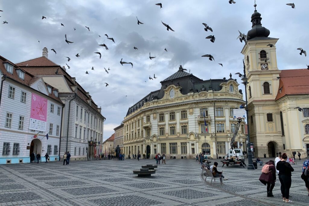 Imaginea prezintă un piață urbană cu clădiri istorice și elegante. Păsări zboară în cerul plimbat de nori, iar oamenii se plimbă, stau pe bancuri sau se întâlnesc. Pe Snapchat, tinerii marocanezi găsesc o varietate de moduri pentru a exprima creativitatea și a-și împărtăși experiențele. Un astfel de piață, cu arhitectura sa frumoasă și atmosfera ei dinamică, ar putea fi un subiect popular pentru storie - ul orar sau filtre creativ pe Snapchat. Tinerii marocanezi pot folosi aplicația pentru a face din acest tip de locuri un spectacol, atragând atenția asupra vieții publice locale și a frumuseții arhitecturale, promovând astfel Marocul și cultura sa printr-un prism modern de comunicare.